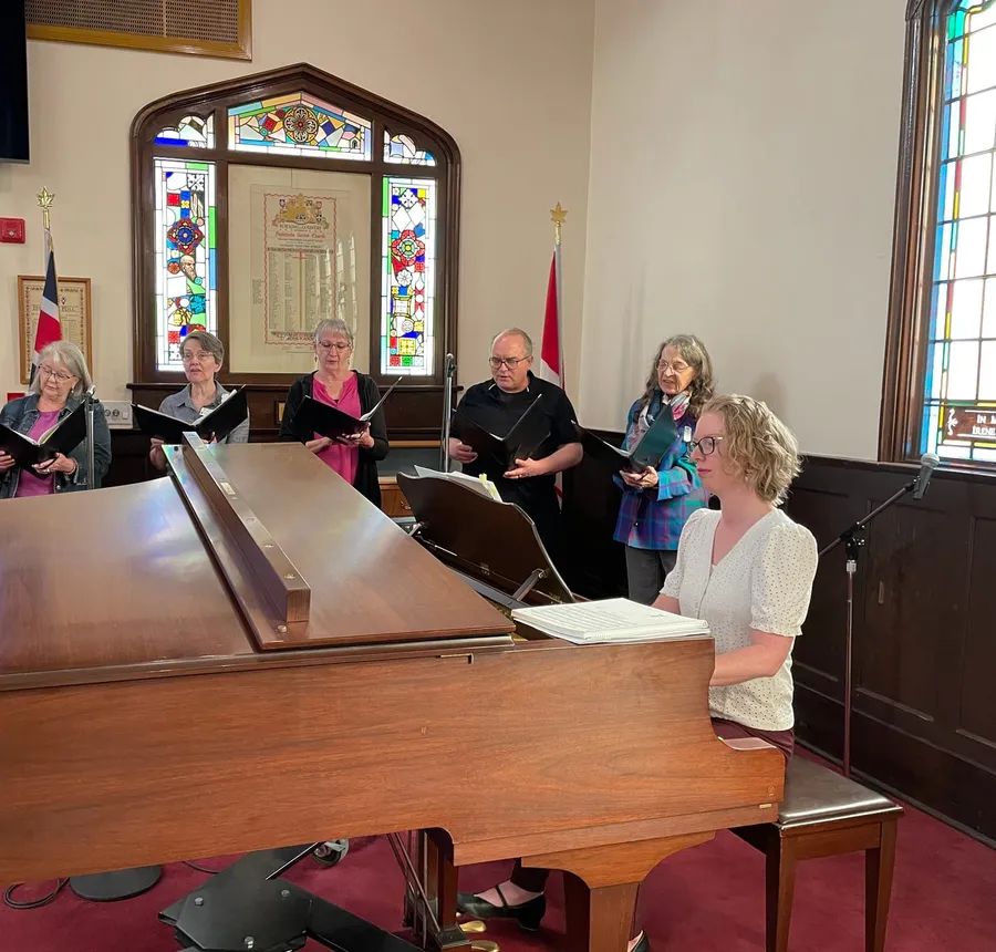The choir singing around the grand piano before a service, the memorial window behind them