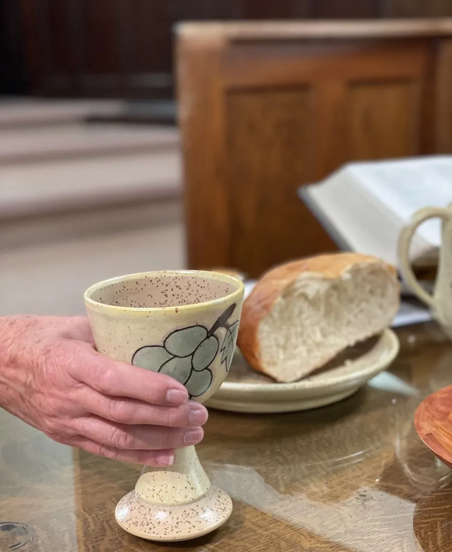 Bread, a pottery chalice, and an open Bible on the communion table, a hand resting on the cup
