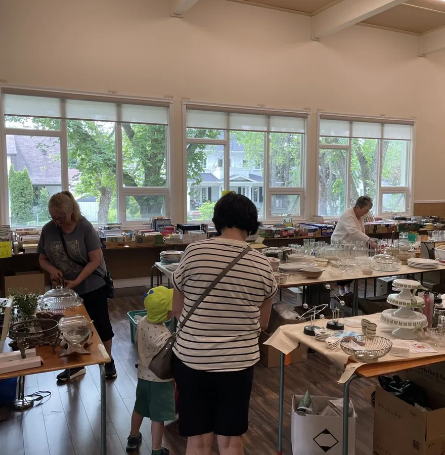 Shoppers browsing tables of glassware and household finds at the community sale, sunlit windows behind
