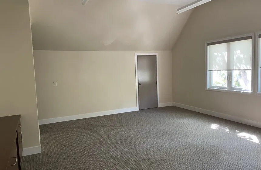 A bright office space with a large window, sloped ceiling, and fresh carpet